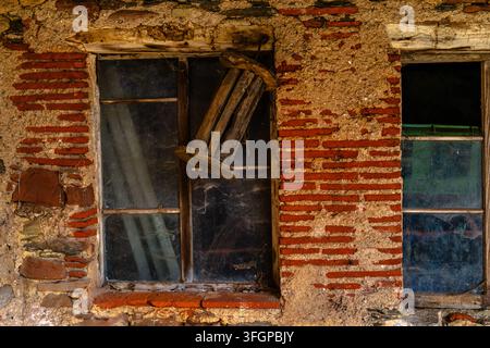 Two old windows with wooden frames set into a deteriorated brick and plaster wall, with a weathered wooden chair visible across one opening. Stock Photo