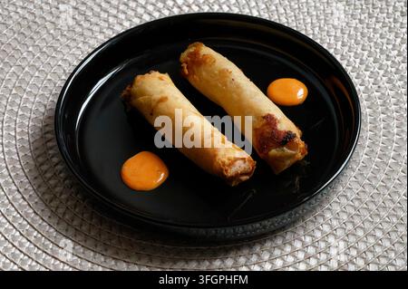 Two golden fried spring rolls served with drops of orange chili sauce on a sleek black plate. Asian appetizer concept. Stock Photo