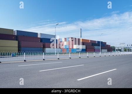 An empty asphalt road with white container yard; stacked freight containers stand with a cable-stayed bridge in the distance under a bright blue sky, background of modern trade and logistics. ? Stock Photo