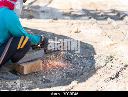 Metalworker crouching down while using a gas cutting torch to slice thick structural steel with sparks spraying on the ground. Stock Photo