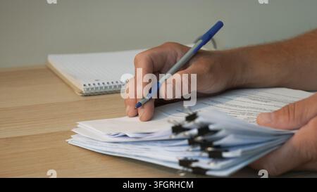 Man holding pen and signing business contract documents on a wooden office table with stacks of paperwork. Stock Photo