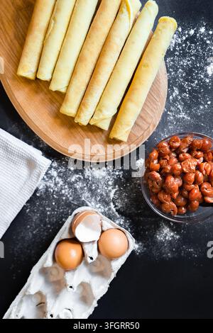 Rolls of tortillas and a bowl of cooked beans are placed on a wooden board. Eggs sit in a carton nearby. Flour is scattered on the counter. Stock Photo