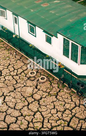 Vertical high angle view of a white and green houseboat grounded on deeply cracked, dry riverbed soil. Selective focus. Stock Photo