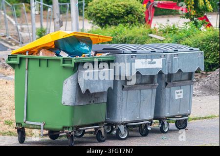 Overflowing green trash bin with yellow lid beside large metal recycling dumpsters outdoors. Stock Photo