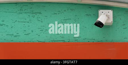 A modern white surveillance camera installed on a vibrant teal stucco wall, providing safety and monitoring in an urban setting with colorful architec Stock Photo