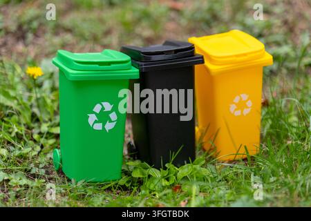 Three recycling bins in green, black, and yellow stand outdoors on grass. Stock Photo