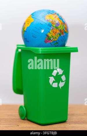 A colorful globe rests inside a green recycling bin on a wooden surface. Stock Photo