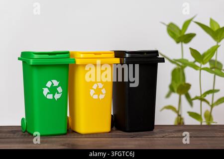 Three colorful recycling bins stand on a wooden table with a blurred plant in the background. Stock Photo