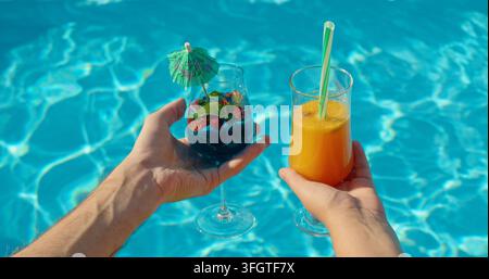 Two hands are shown holding elegant cocktail glasses filled with refreshing drinks, featuring fruit and soda. The pool water surface in the background Stock Photo