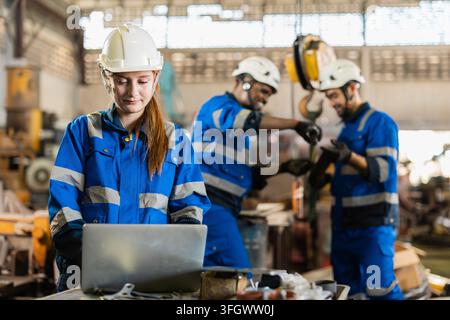 Female engineer wearing safety helmet using laptop computer inside old heavy machinery warehouse storage facility to check technical inventory and mai Stock Photo