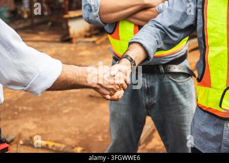 close up construction workers engineers wearing safety vests hardhats shaking hands agreement partnership handshake outdoor building construction site Stock Photo