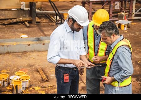 group diverse construction workers engineers hardhats reflective safety vests looking digital tablet discussing planning blueprint building developmen Stock Photo