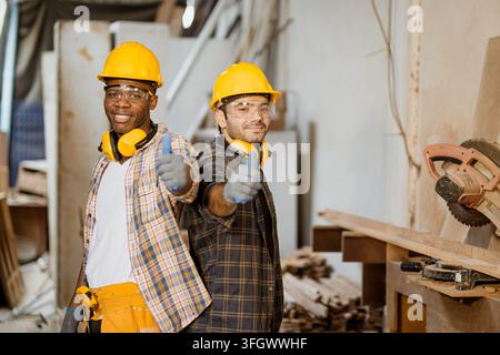 Two Male Carpenters Workers Showing Thumbs Up Gesture Inside Woodwork Factory Workshop, Multiethnic Worker Smiling Teamwork Success Agreement Hard Hat Stock Photo