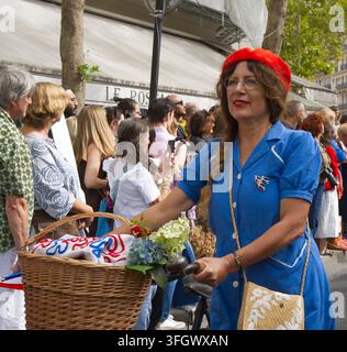 A woman dressed in a vintage blue dress and bright red beret pushing a bicycle with a flower-filled wicker basket during a parade in Paris, France. Stock Photo