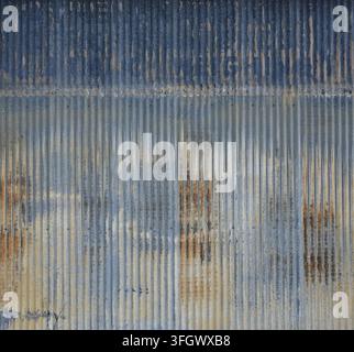 Blue tinted corrugated galvanized metal surface showing a dark blue upper band, battered tan paint remnants, faded blue lower panels, rust Stock Photo