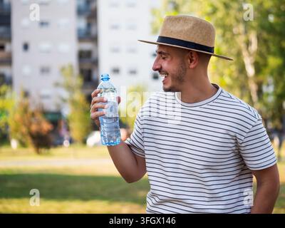 Young happy adult man drinks water in park on sunny summer day. Stock Photo