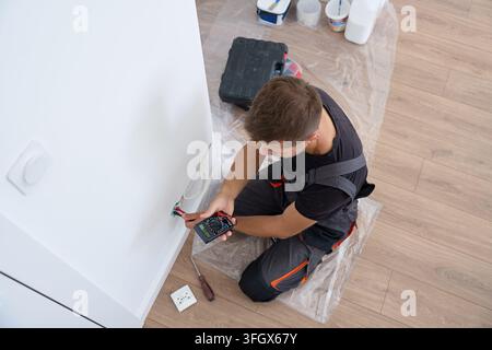 Electrician checks wires for a wall socket with a digital multimeter. Electrical work, installing power outlets and home renovation. Stock Photo
