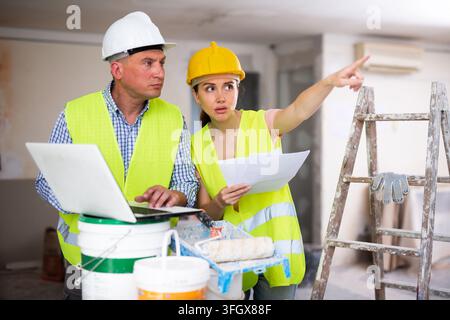 Portrait of woman architect and foreman holding blueprints and using laptop while discussing working process in house being renovated Stock Photo