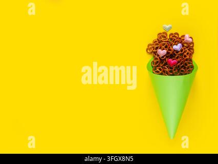 Mini pretzels and small colorful hearts in light green paper cone lie on yellow background. Pretzel sunday concept banner Stock Photo