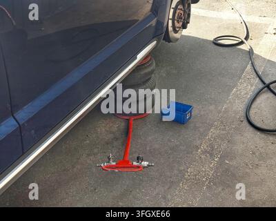 Blue car lifted with a red hydraulic jack and stacked tires underneath, with a blue plastic crate and black hose on the concrete ground beside it. Stock Photo