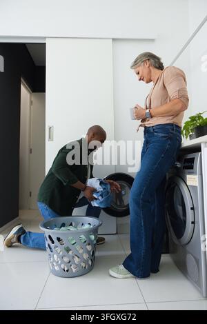 Diverse couple kneeling and standing in laundry room loading clothes into front-load washer Stock Photo