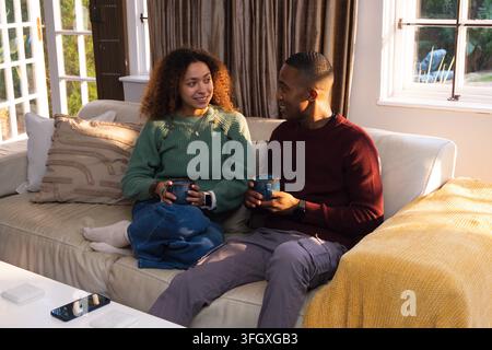 Diverse couple sitting on beige sofa at home, holding blue mugs, phone on table, yellow throw Stock Photo