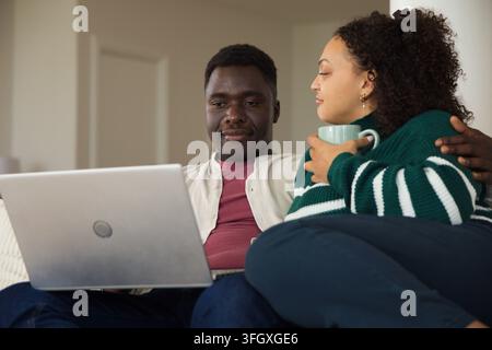 African American couple sitting on light sofa at home, sharing silver laptop and light blue mug Stock Photo