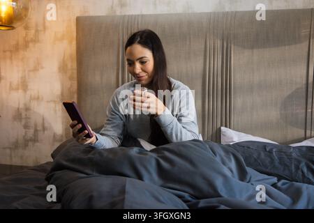 Woman sitting up in bed holding smartphone in left hand and glass in right under lamp Stock Photo