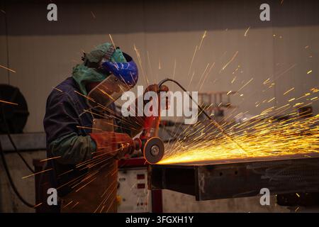 View of a worker wearing protective gear using an angle grinder on a metal beam, creating a shower of bright sparks in a workshop in Milan, Lombardy, Stock Photo