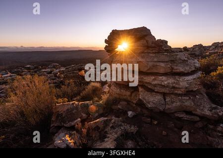 View of a natural rock formation with a sunburst shining through a hole in the stone, set against a vast rocky landscape during golden hour in South A Stock Photo