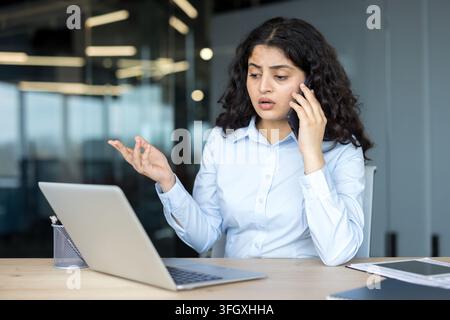 Businesswoman Talking On Smartphone And Working On Laptop In Office, Professional Employee Managing Corporate Communication And Financial Analysis At Desk. Confident Indian Manager Analyzing. Stock Photo