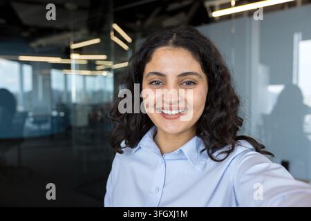 Confident Businesswoman Taking A Selfie In A Modern Corporate Office, Smiling Professional Employee Working In A Bright Workplace Environment. Portrait Of A Successful Manager Using A Smartphone. Stock Photo