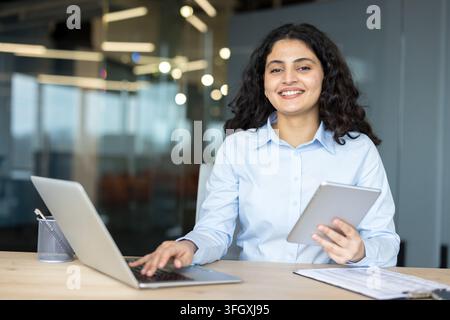 Confident Businesswoman Working On Laptop And Tablet In A Corporate Office Setting For Financial Analysis And Strategic Planning. The Professional Female Employee Is Using Digital Technology At Her. Stock Photo