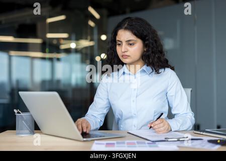 Focused Businesswoman Working On Laptop And Writing Documents At Desk In Corporate Office. Professional Manager Analyzing Financial Analysis And Planning Strategy While Using Laptop And Writing. Stock Photo