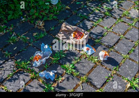 Iserlohn, Germany - July 13, 2026: Discarded Burger King paper bag and food wrappers littering a paved sidewalk with weeds. Stock Photo