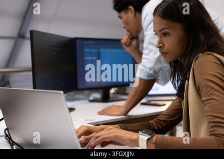 Diverse coworkers in business wear coding at desk, woman typing laptop while man viewing monitors Stock Photo