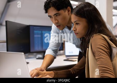 Diverse coworkers reviewing code on laptop at desk with dual monitors and phone, wearing shirt Stock Photo