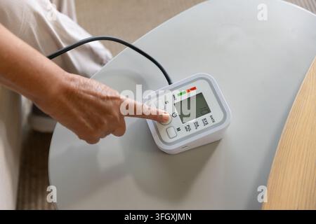 Senior African woman pressing start on BP-monitor, cuff tubing on side-table at home in light pants Stock Photo