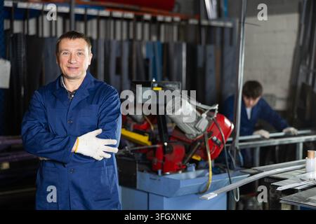 Portrait of confident smiling experienced male craftsman, wearing blue work coat and gloves, standing with arms crossed in industrial metalworking wor Stock Photo
