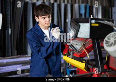 Focused young workman in blue work jacket and gloves working on metal profile cutting machine in professional processing workshop Stock Photo