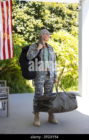 Female soldier standing on porch carrying olive duffel and black pack, US flag visible nearby Stock Photo