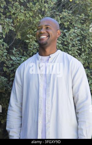 Adult African man standing and smiling by variegated shrubs wearing pale button-up shirt Stock Photo