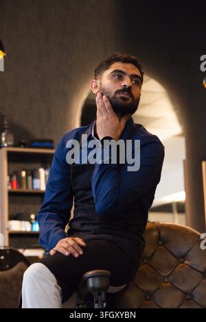 Man sitting in tufted chair touching beard, wearing dark blue shirt and black apron at barbershop Stock Photo