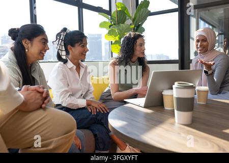 Diverse female coworkers collaborating around table in office lounge using laptop and coffee cups Stock Photo