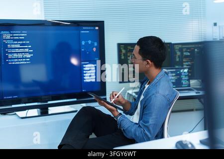 Focused developer reviewing programming code on multiple screens while taking notes on a tablet, representing software development, cybersecurity, dig Stock Photo
