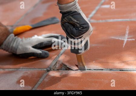 Worker wearing protective gloves removes damaged tiled flooring with a hand chisel during floor renovation Stock Photo