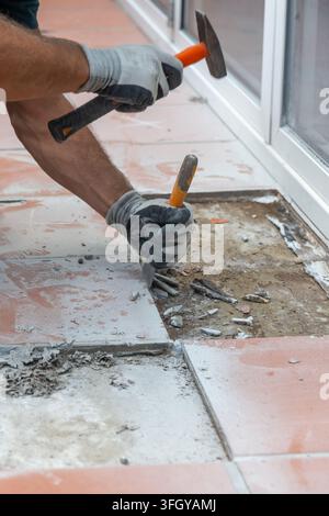 Worker wearing protective gloves removes old ceramic floor tiles using a hammer and chisel during renovation Stock Photo
