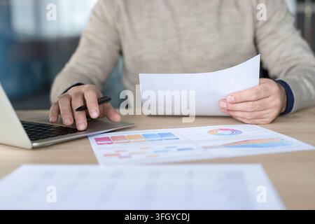 Businessman Working On Financial Analysis With Laptop And Documents At Desk In Office. Professional Manager Reading Paper Report And Using Computer For Strategic Planning In Corporate Workplace. Stock Photo