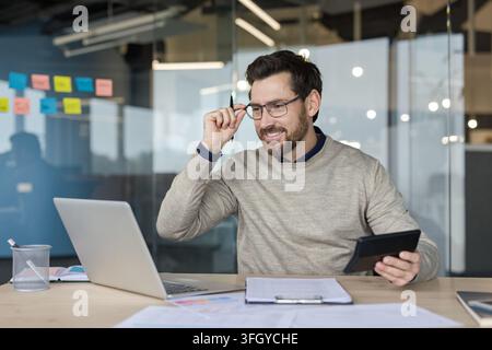 Businessman Working On Laptop And Analyzing Documents At Desk In Corporate Office. Professional Manager Reading Financial Reports And Planning Strategy While Using Computer In Modern Workplace. Stock Photo