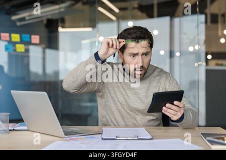 Stressed Businessman Looking At Tablet In Office, Shocked Manager Analyzing Financial Documents And Laptop Data. Professional Executive Working At Desk With Paper Documents And Computer While Feeling. Stock Photo